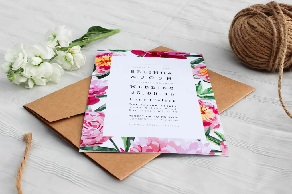 Wedding invite showcasing it's design of pink gerbera flowers and leaves surrounding the border with invite details are displayed in the middle. The invite is positioned on a white clothed table laying on top of a brown envelope surrounded by a flower stem and a ball of twin.
